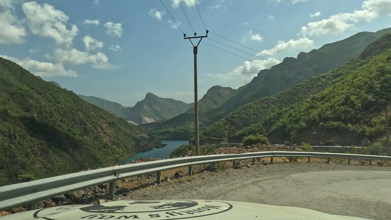 Bocht in de SH25 met zicht op Komani Lake en metalen vangrail langs de weg.