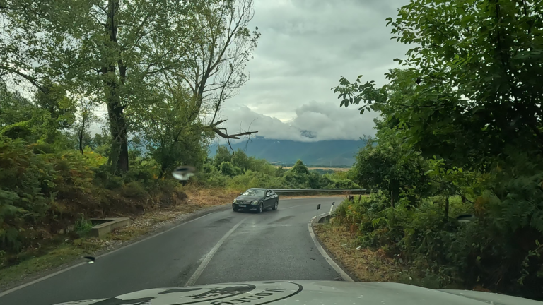 Bochtige weg: Rruga Azem Hajdari, met bomen en uitzicht op een vallei met donkere wolken boven de bergen.
