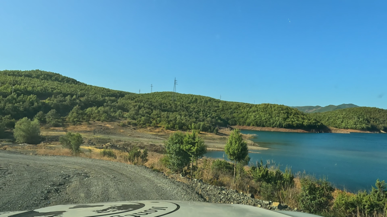 Gravelweg langs het Qafë Mollë-stuwmeer met bomen en strandachtige oever.