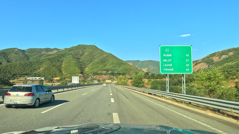 Snelweg A1 in Albanië met passerende auto en groen bord langs bergachtig landschap.
