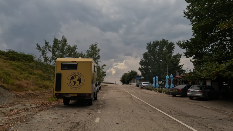 Camper Monty geparkeerd langs SH3 (Albanië) bij restaurant Ismet met blauwe parasols, bomen en geparkeerde auto’s onder een grijze lucht.