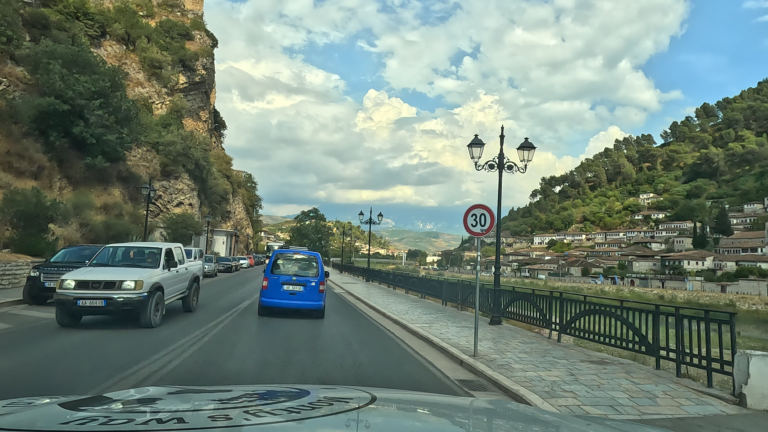 Rivier langs SH3 in Berat met stoep, hek en huizen op een heuvel aan de overkant.