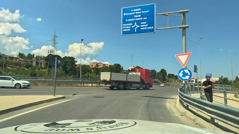 Asfaltweg met verkeersborden boven de rijbaan en een vrachtwagen die op de voorrangsweg rijdt.