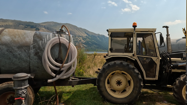 De tractor en waterwagen van Gennaro staan bij het waterpunt aan Lago Matese, met het meer en gebergte tussen beide voertuigen.