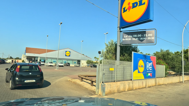 Ingang van Lidl met groot blauw logo en parkeerterrein achter het hek in San Vito
