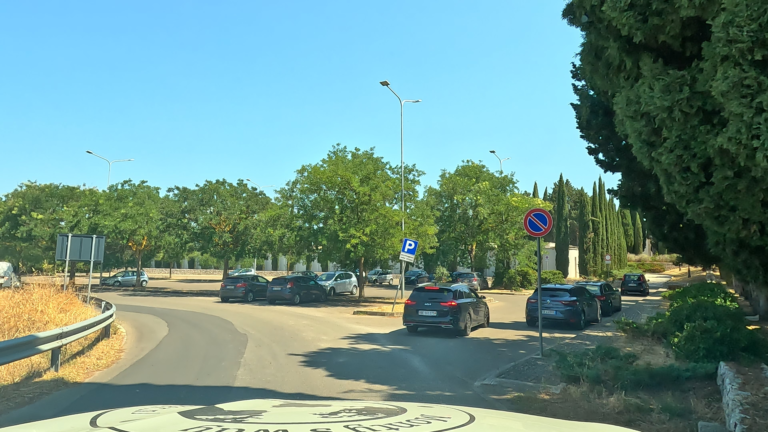 Parkeerplaats bij Cimitero Comunale Noci met auto's tussen olijfbomen en cipressen langs de muur