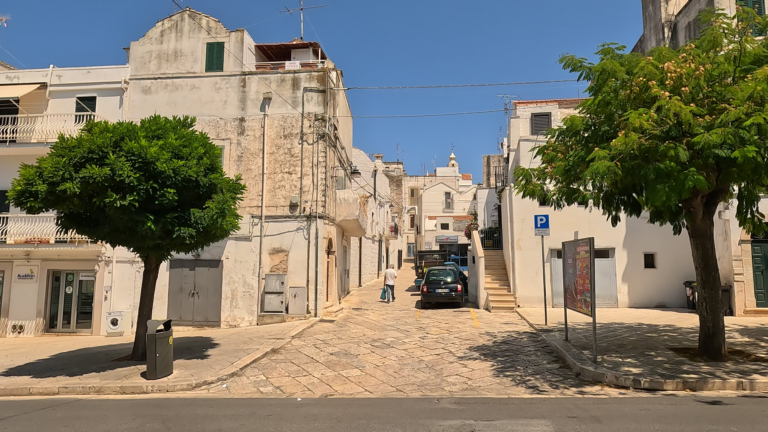 • Tijdens de rit richting Matera zie ik typische witte huizen van Noci langs een straat met basole stenen, bomen en voetganger in het centrum