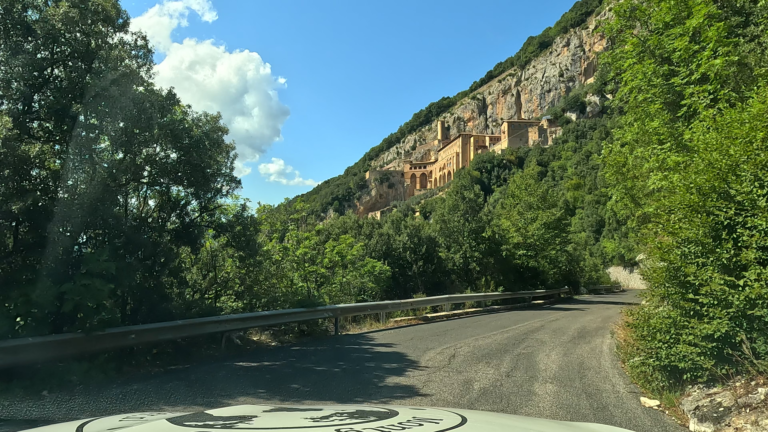 Eerste blik op Monastero di San Benedetto boven de bomen tegen de rotswand langs de SP45A tijdens de route naar Subiaco.